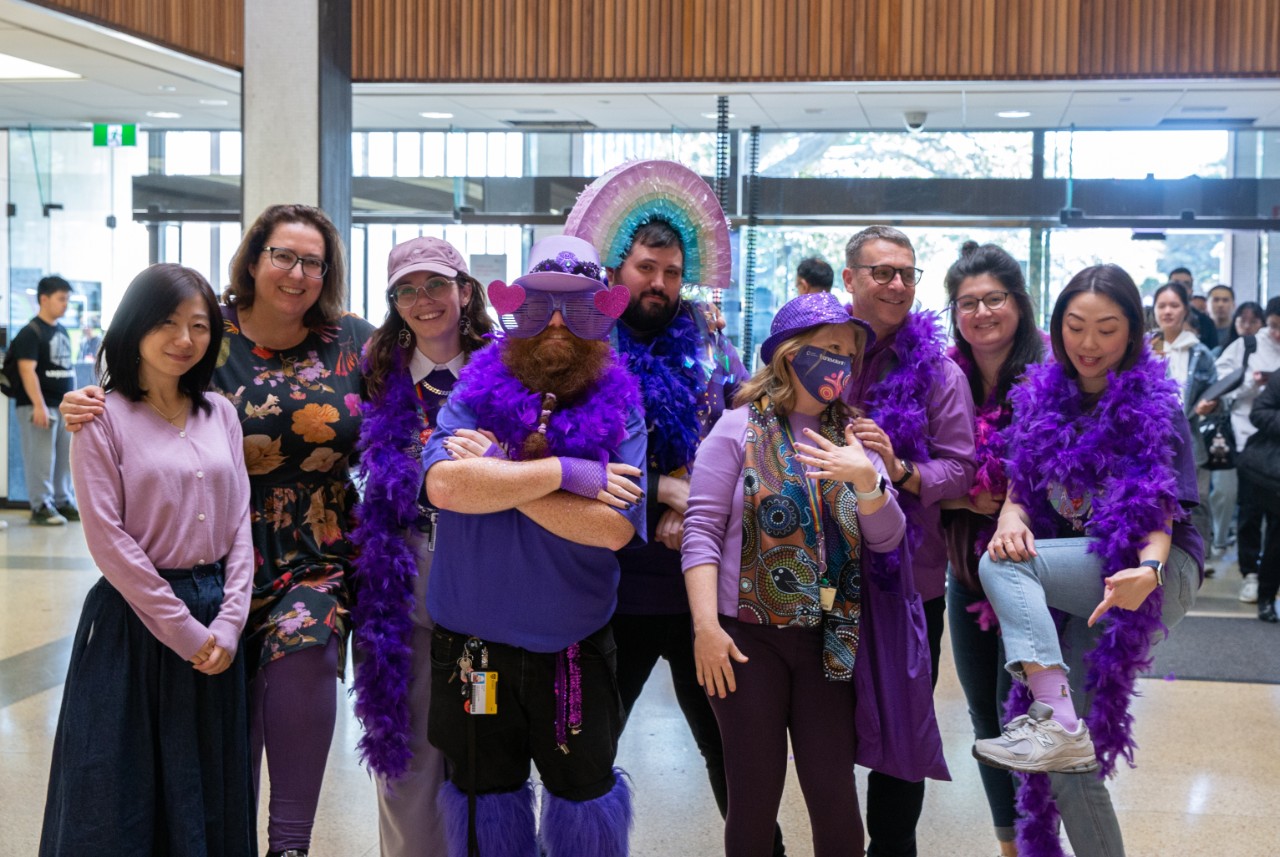 A photograph of a group of Library staff members wearing purple clothing, including extravagant flourishes like feather boas and papier mache hats