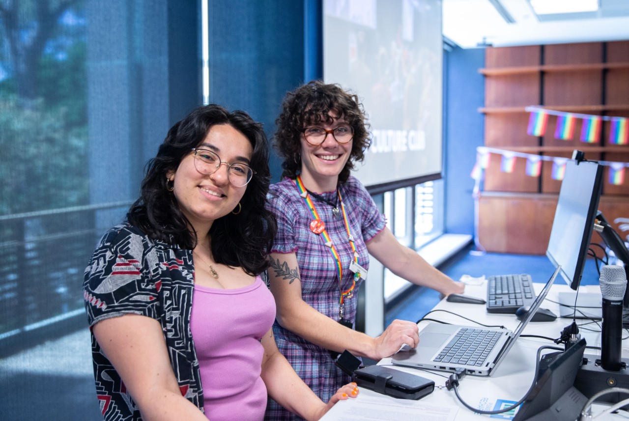 A photograph of two smiling women standing at a lectern in front of a computer. One of them is wearing a rainbow University of Sydney lanyard with a "she/her" pronouns badge. In the background is rainbow pride bunting.