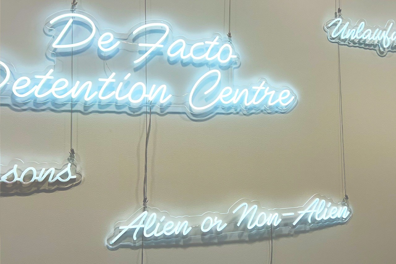 A close up shot of a series of white neon signs, with a focus on the words De Factor Detention Centre and Alien or Non-Alien