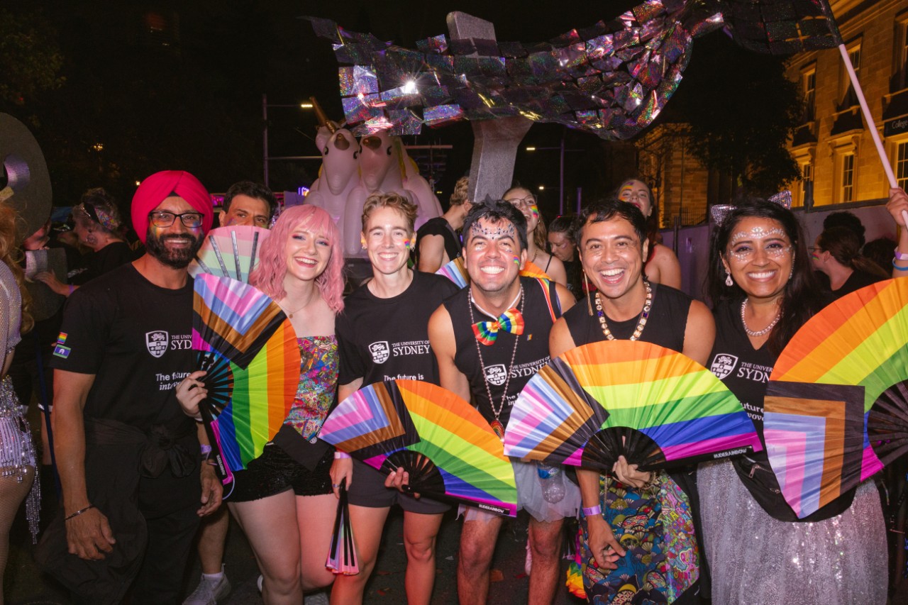 A group of people smiling at the camera. They are wearing matching outfits, with colourful decorations and are holding pride progress flags