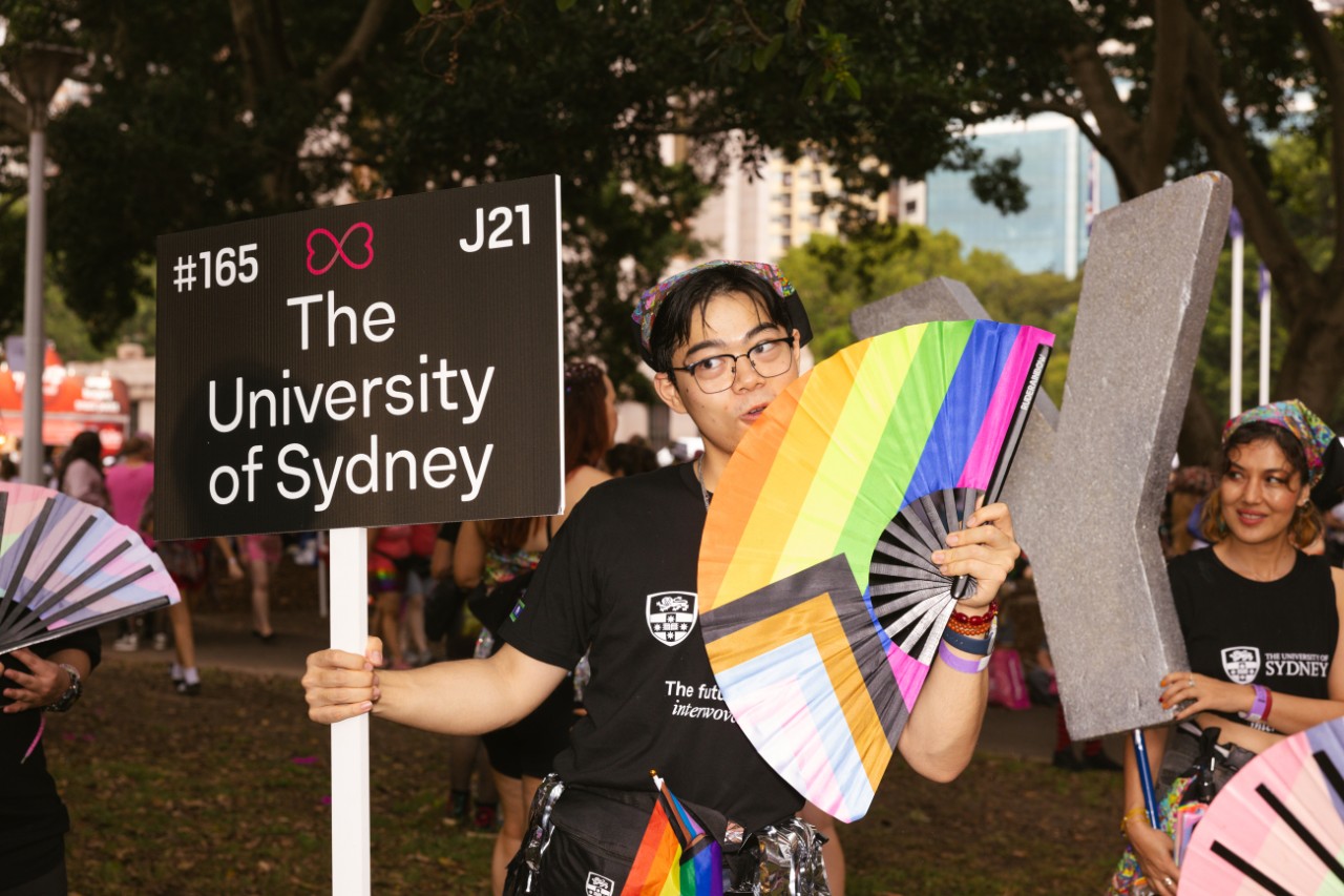 A person holding a fan with the pride progress flag in one hand and a 'The University of Sydney' sign in the other