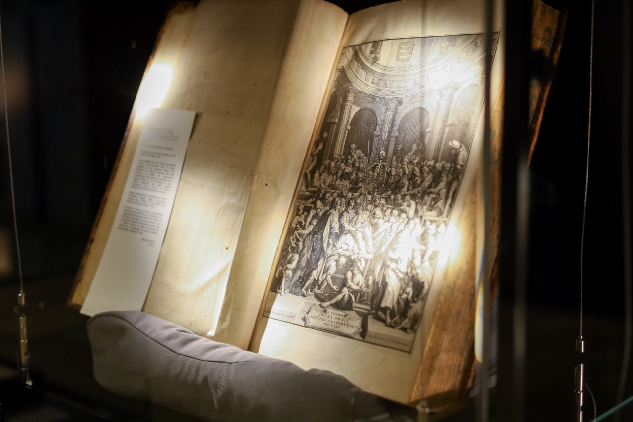 An open book in a glass cabinet, featuring an intricate illustration from the first page of Vesalius' book.