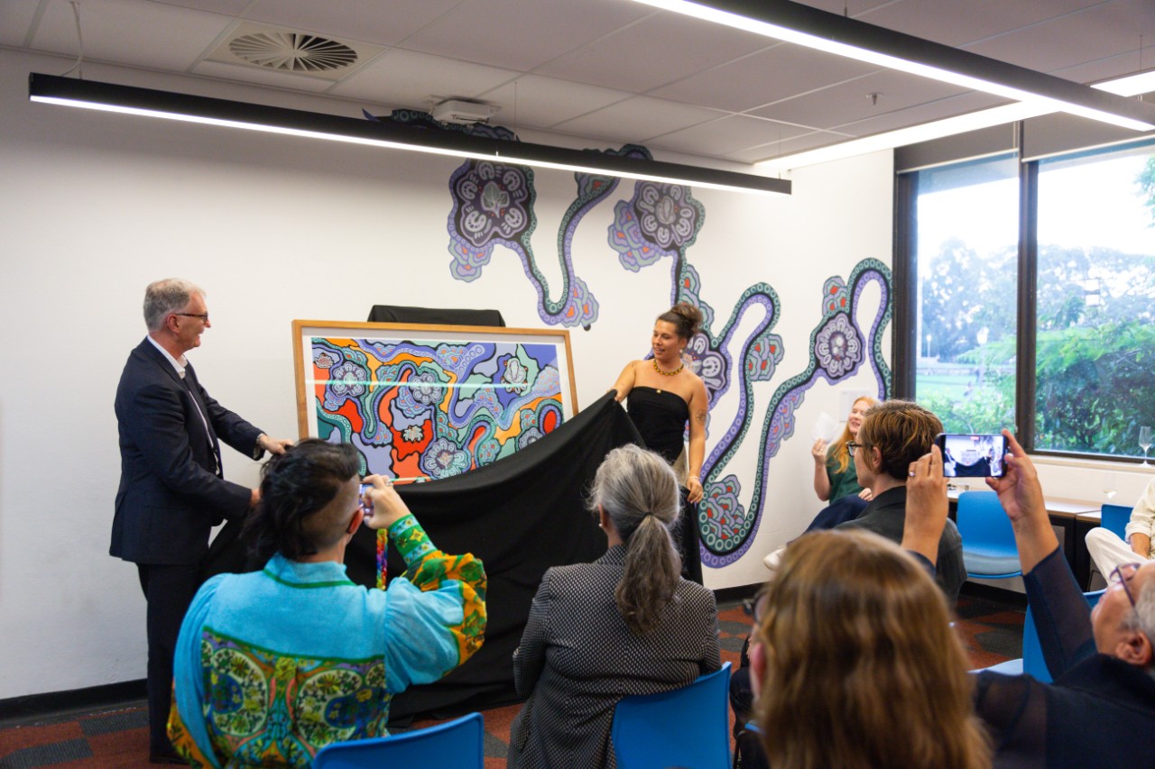Philip Kent and Jessie Waratah removing a black cloth to reveal the artwork Walking on Country displayed on an easel