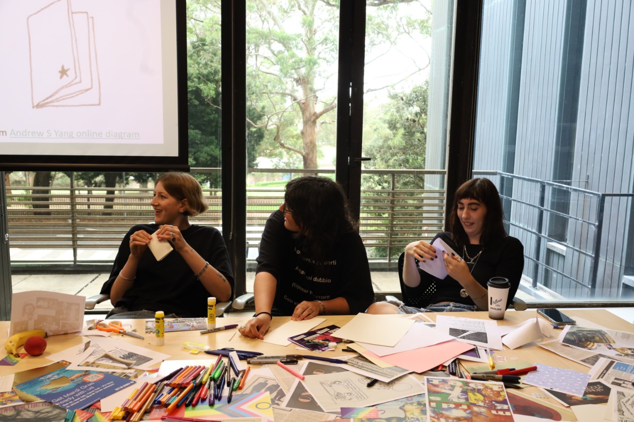 A colour photograph depicting three people smiling while sitting at a table scattered with craft materials