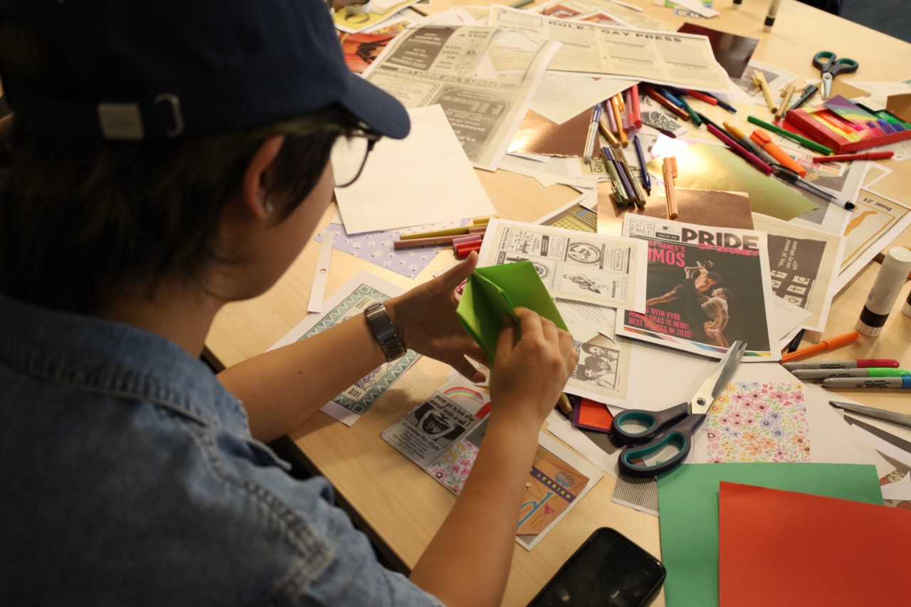 A close up photograph taken over someone's shoulder while they create a zine. The table is scattered with craft materials.