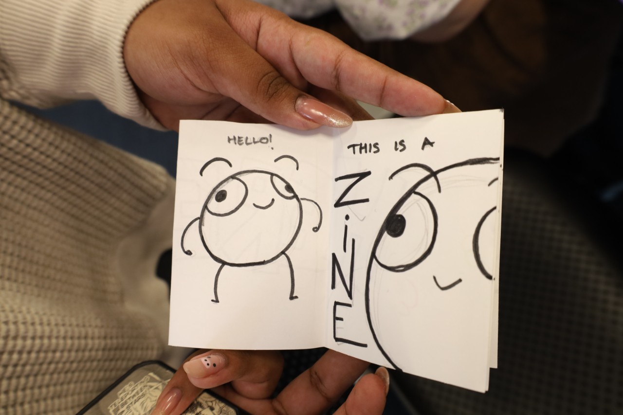 A hand drawn spread from a zine, showing a cartoon figure saying "Hello! This is a ZINE"