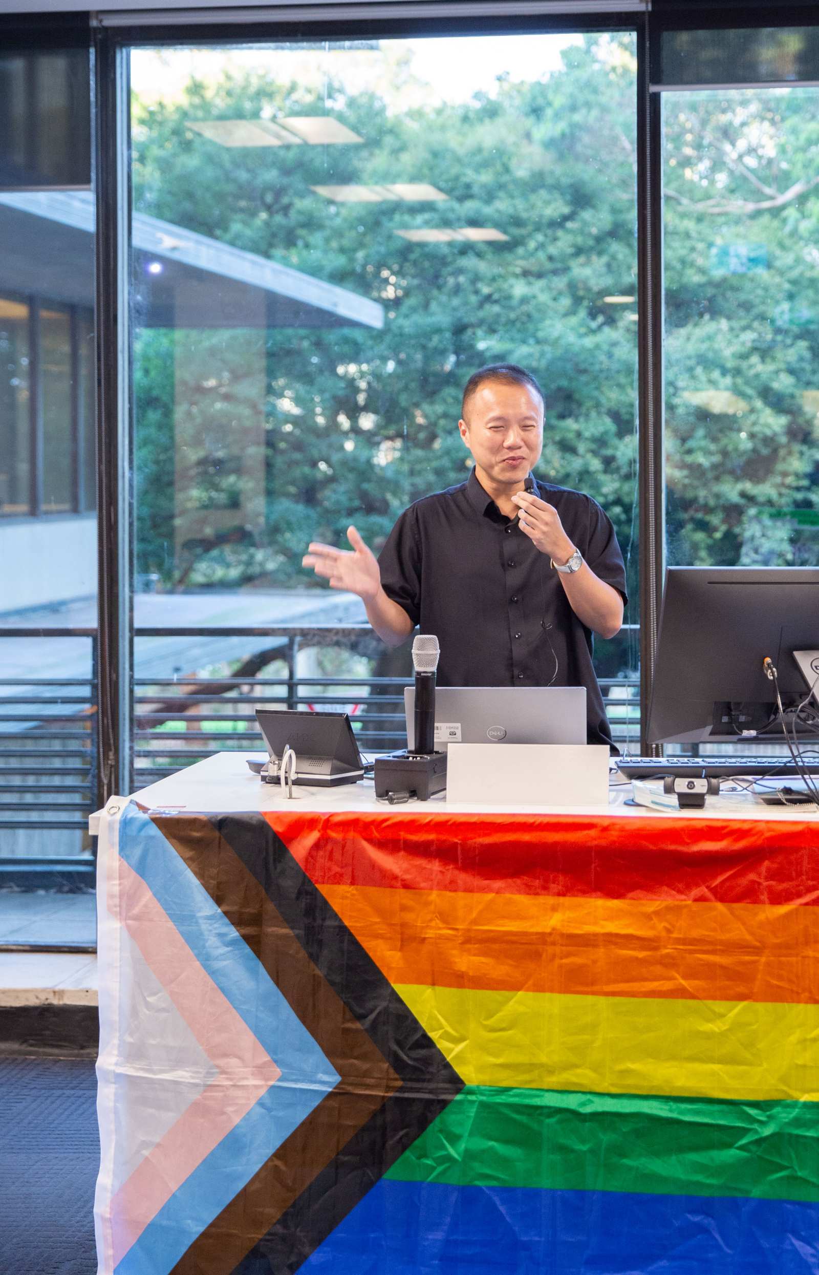 A photograph of Gavin Lee (a person with very short hair wearing a black shirt) presenting at a rainbow flag adorned lectern. He stands in front of a window, beyond which lies a green park.