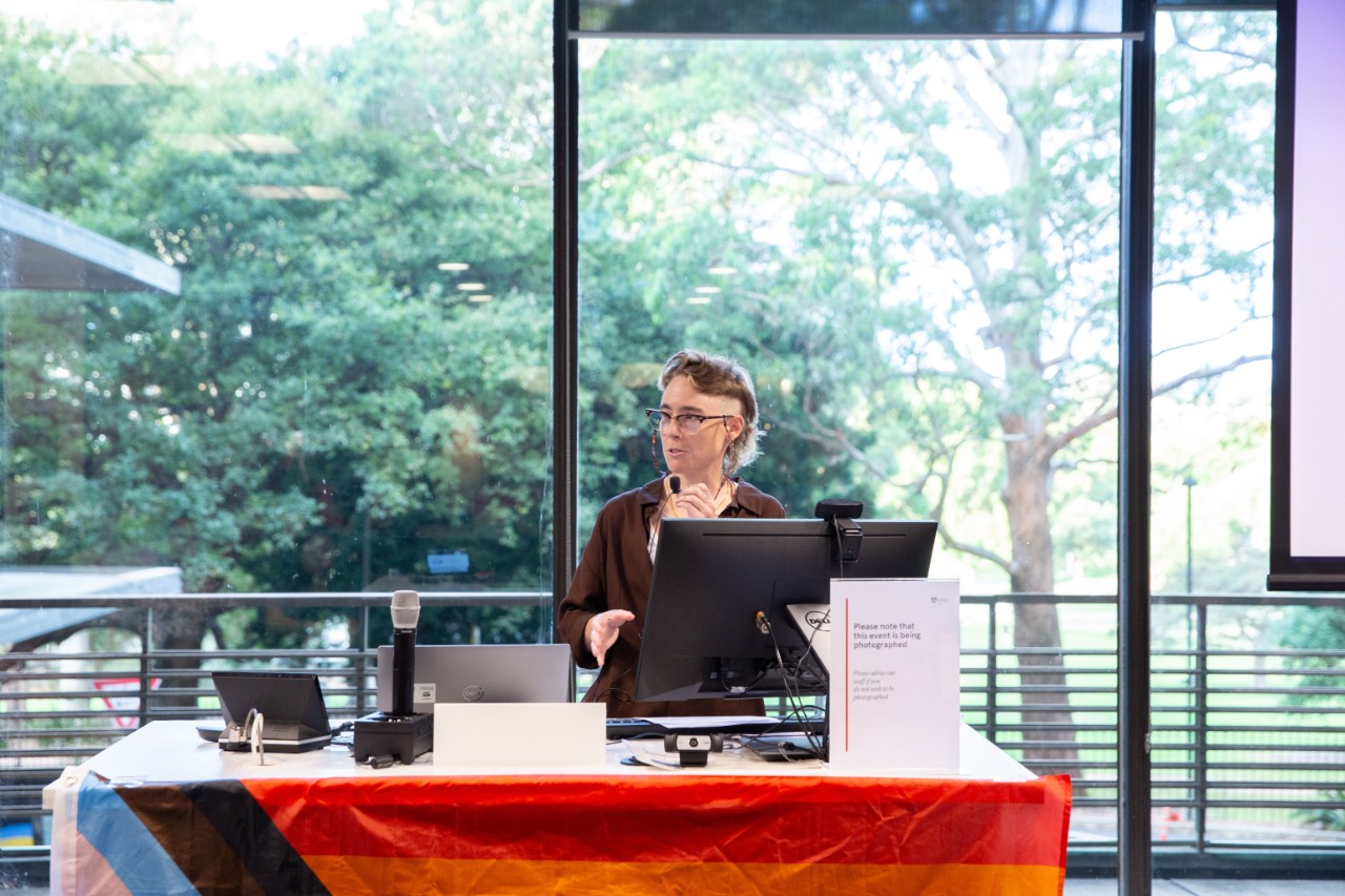 A photograph of Alexandra Pinkham (a person with a mullet and glasses) presenting at a rainbow flag adorned lectern in front of a window, beyond which lies a green park.