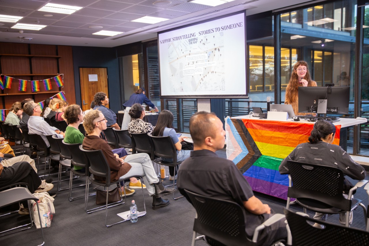 Photo of olivia sutherland (a person with long red hair and glasses) presenting to an attentive audience. Rainbow flags adorn the lectern and walls.