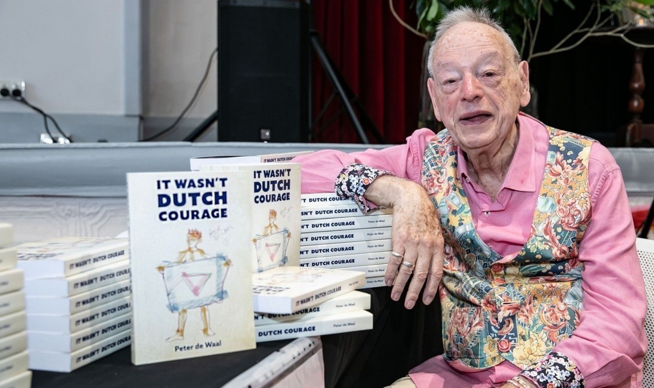 A colour photograph of Peter de Waal (an elderly man wearing a pink shirt and colorful vest) next to several copies of his memoir "It Wasn't Dutch Courage"