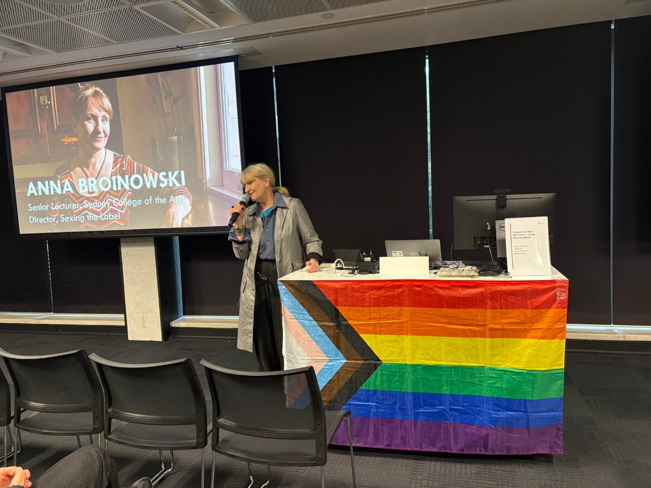 A photograph of Gavin Lee (a person with very short hair wearing a black shirt) presenting at a rainbow flag adorned lectern. He stands in front of a window, beyond which lies a green park.