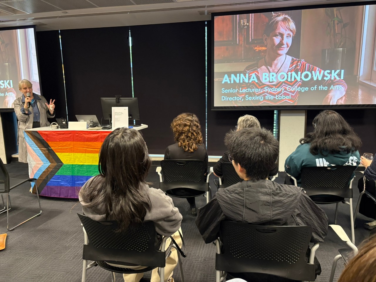 Photo of Anna Broinowski (an older woman) presenting to an audience, a rainbow flag covering the lectern in front of her.