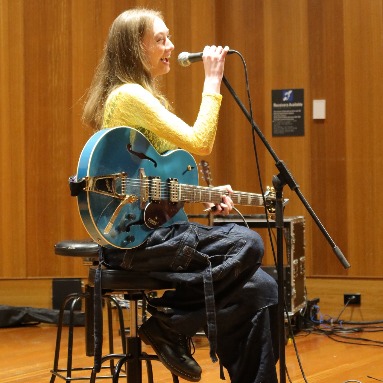 A colour photograph showing Cara Sullivan (a young woman with long hair) speaking into a microphone and holding a blue guitar.