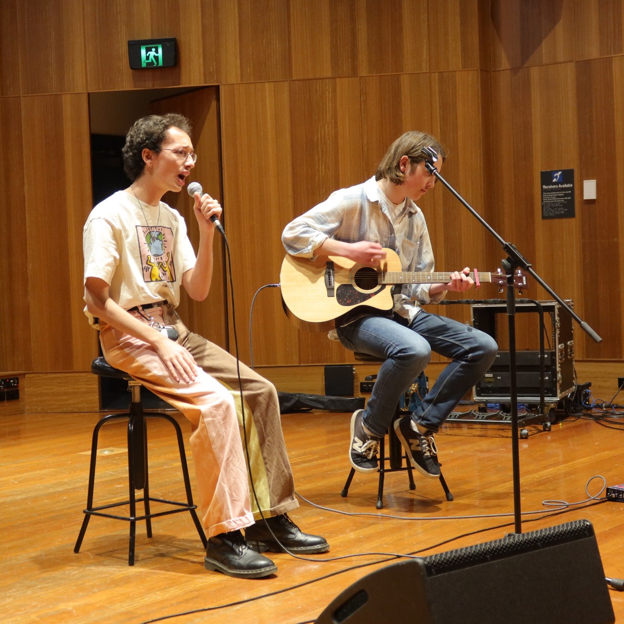 A photograph of two young man performing. In the foreground Samuel Dunnicliff is singing into the microphone. In the background Cameron Spry is playing guitar.