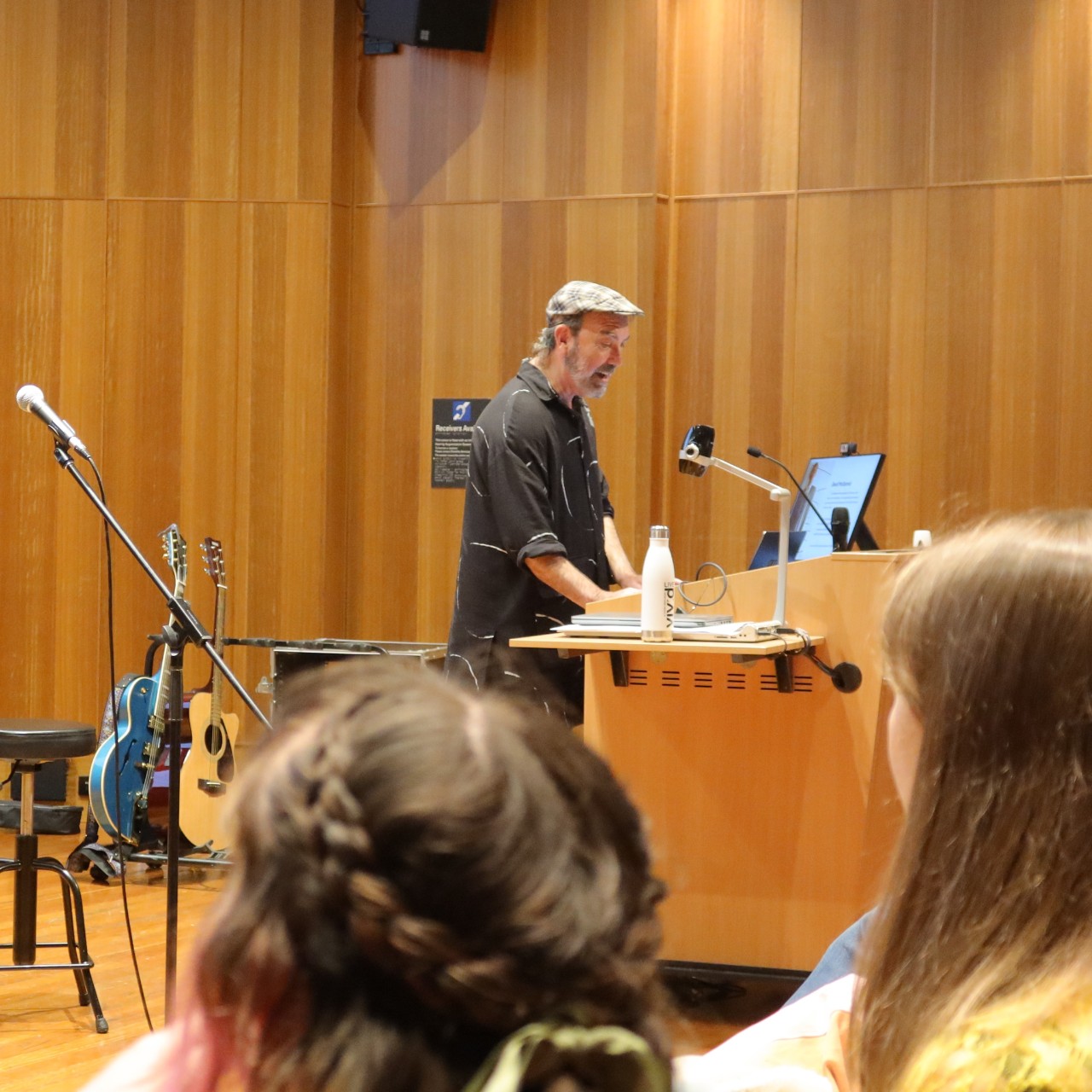 A photograph of Paul Mac (a white man wearing a newsboy cap) presenting to an audience, with guitars in the background