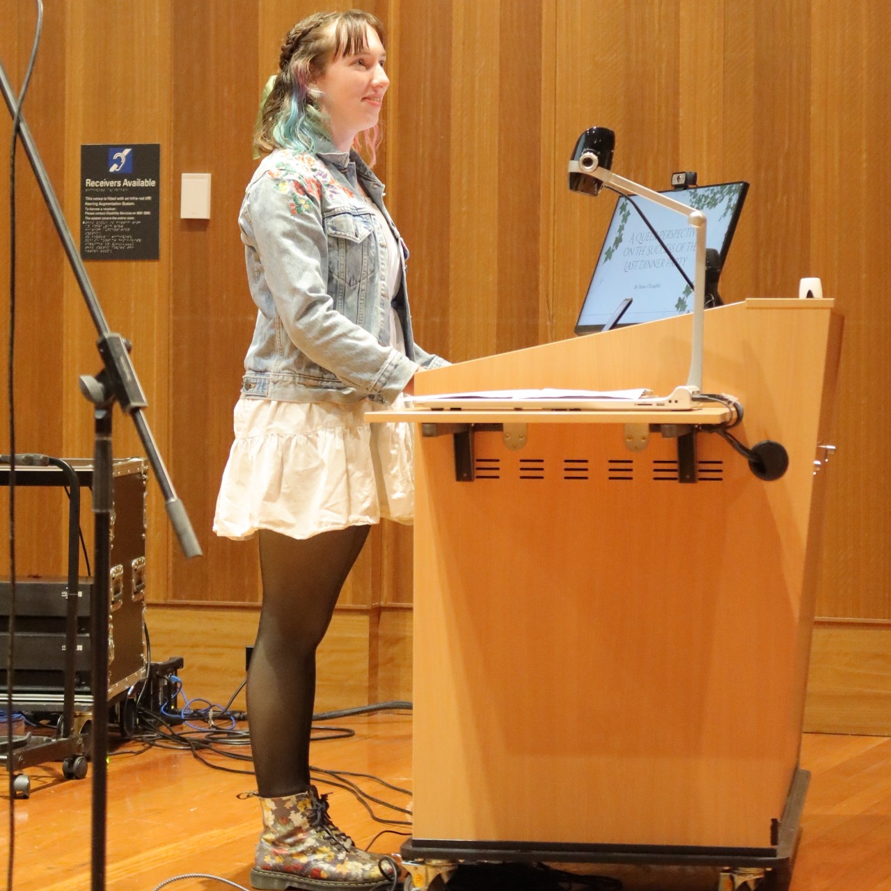 A photograph of Damia O'Loughlin (a young white woman with blue and pink dip-dyed hair) speaking at a lectern. 