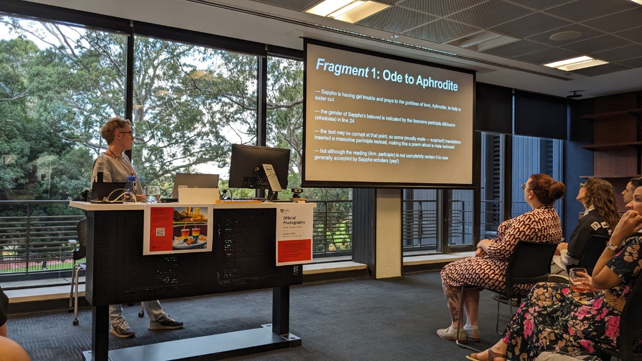 A photograph showing a fair-skinned person with short hair presenting at a lectern. In the background is a projector screen showing a presentation slide with the heading "Fragment 1: Ode to Aphrodite"