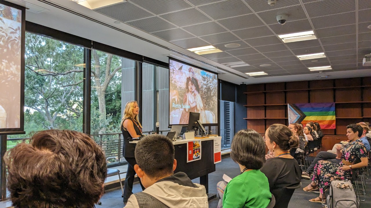 A photograph showing a fair-skinned person with long blonde hair presenting at a lectern to an attentive audience. In the background is a LGBTQIA+  pride progress flag and a projector screen showing a presentation slide that reads "Poetry Saloon: open mic"