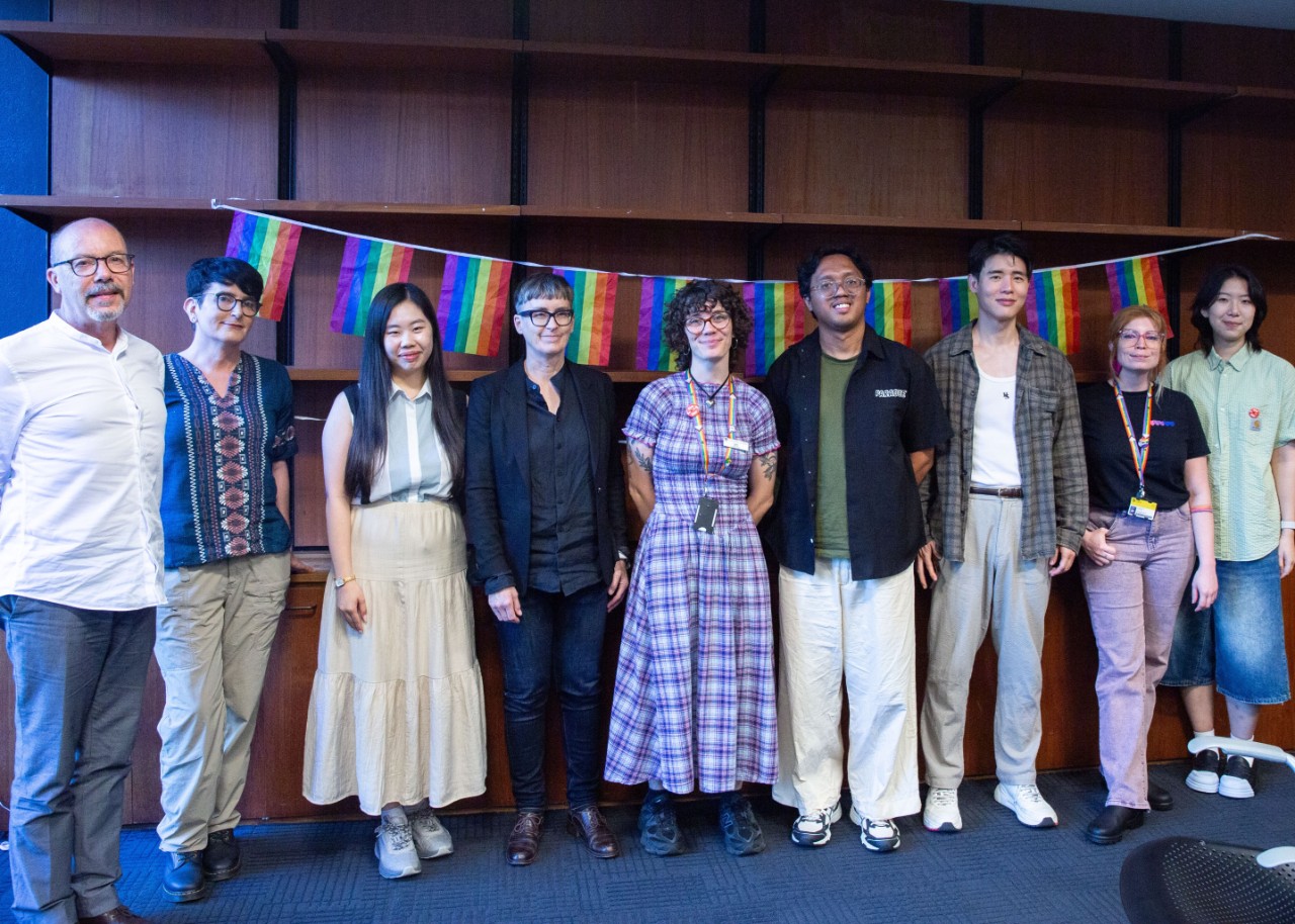 A colour photograph showing a group of 9 people (the presenters and MCs of Pride Culture Club) standing in front of rainbow flags and smiling
