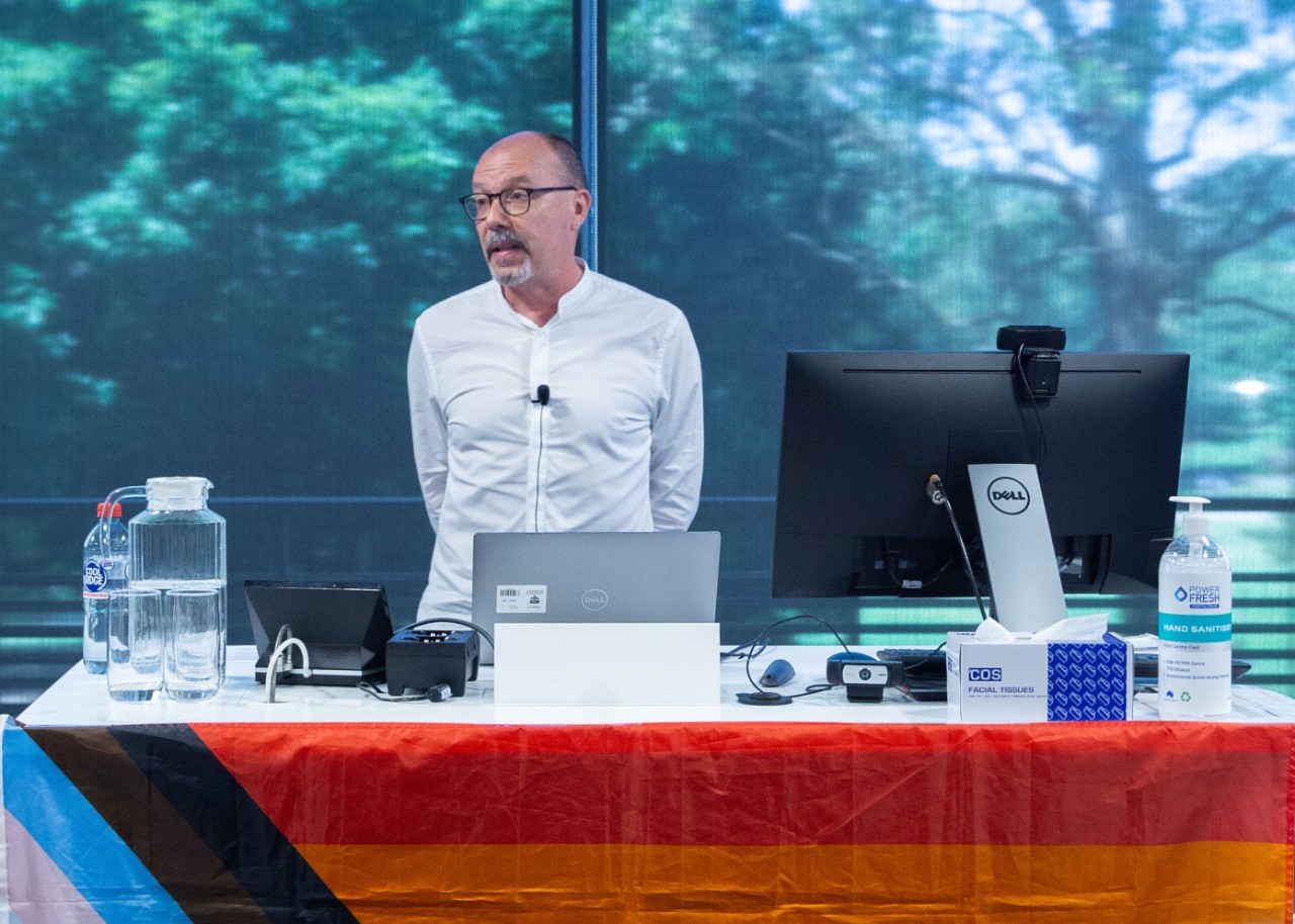 Photo of Dr Allen George presenting behind a rainbow-flag-covered lectern