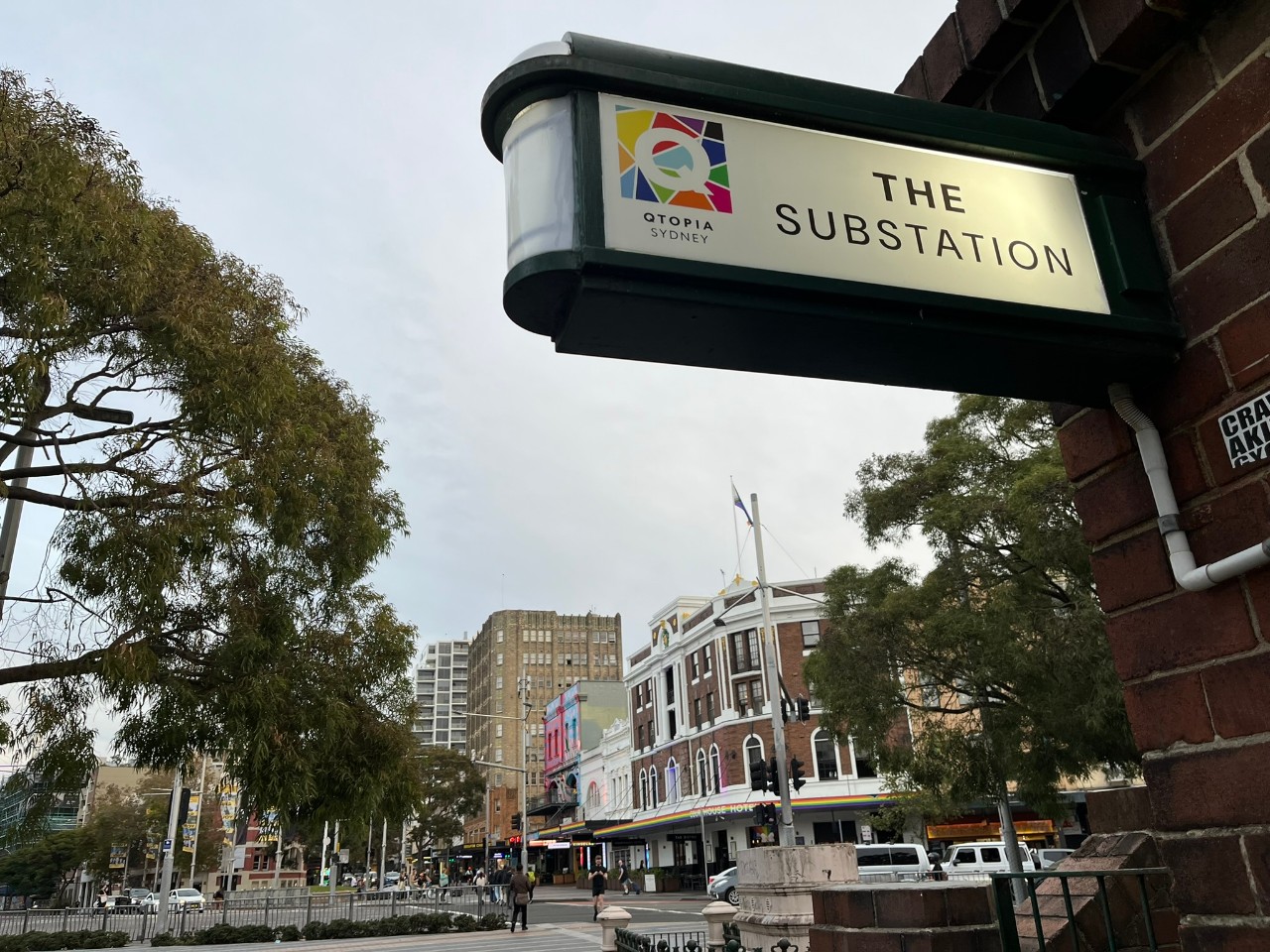 A photograph looking out across Taylor Square from Qtopia, in Darlinghurst, Sydney. In the foreground is a sign that reads "Qtopia Sydney: The Substation" and in the background are trees, a busy city street, and buildings. The Courthouse Hotel's sign is rainbow to indicate that it is a safe space for the LGBTQIA+ community.