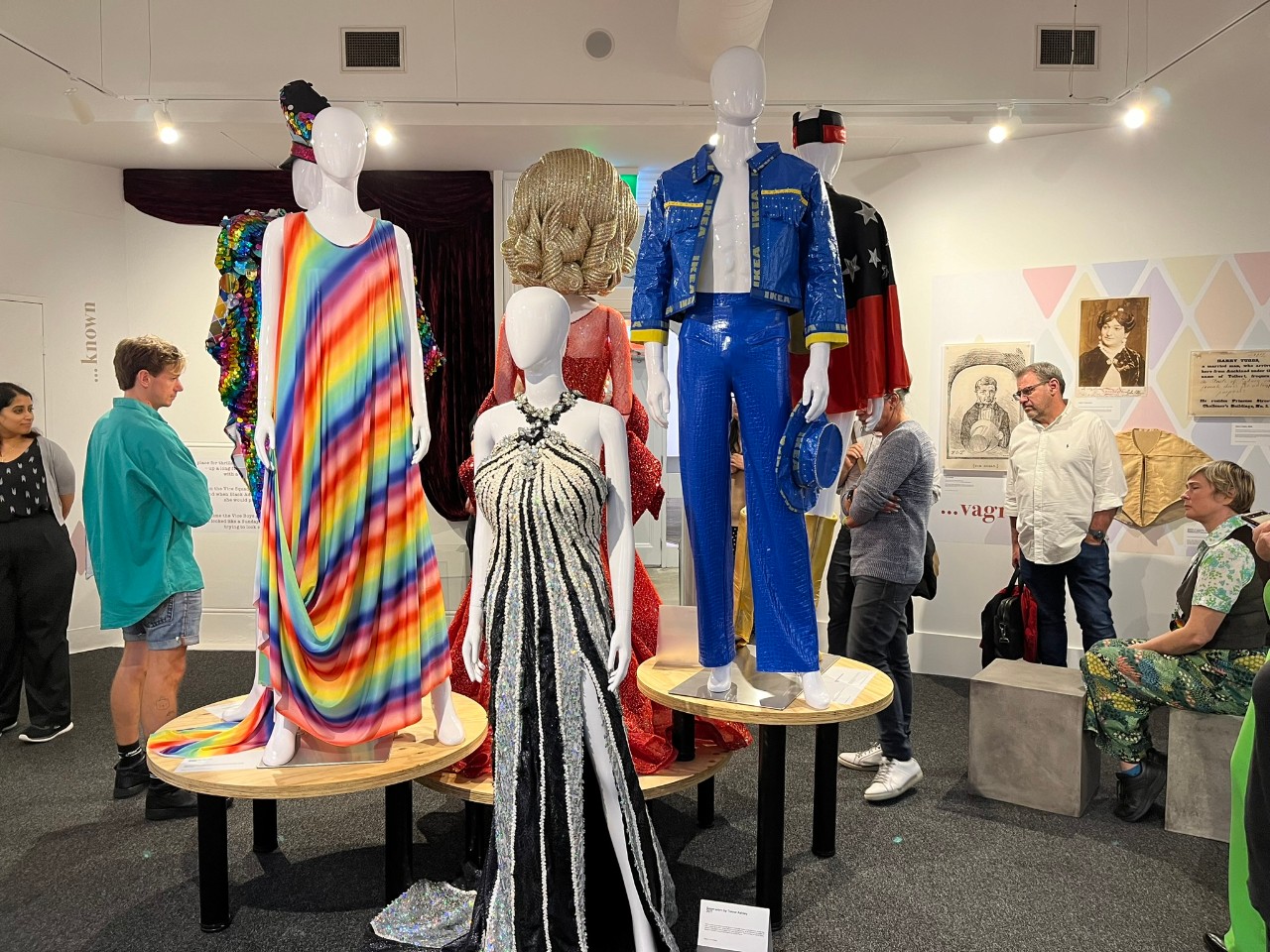 A photograph of an exhibition featuring 6 mannequins in brightly coloured and sequinned drag costumes. Of the three which facing the camera, are the one on the left is wearing a loose fitting rainbow dress, the one in the middle is wearing a black and white sequinned evening gown, and the one on the right is wearing a blue jacket and pants made out of IKEA bags. People are standing around the display looking at it and other things in the room.