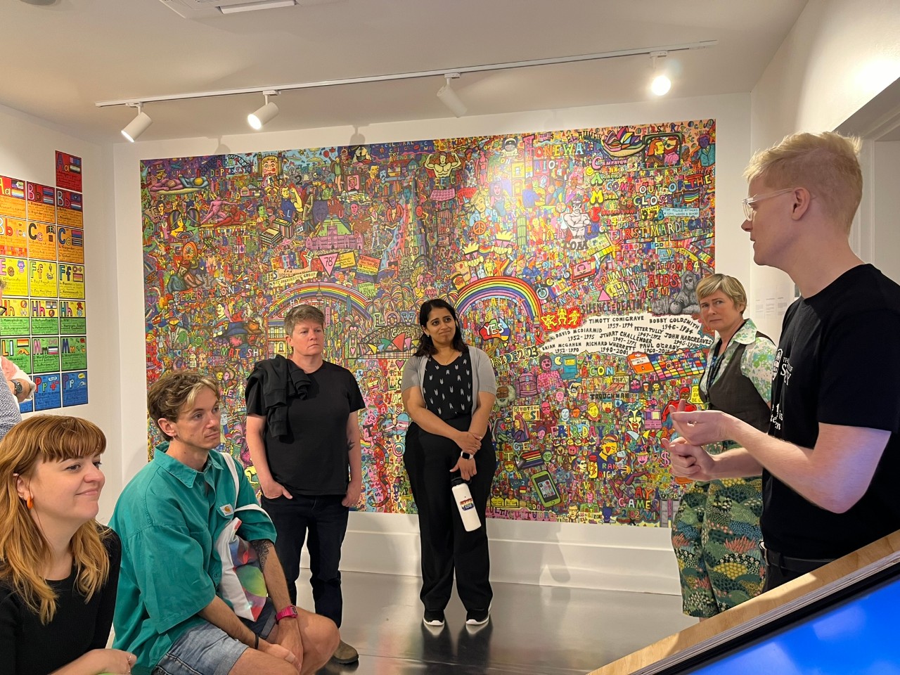 A photograph showing a group of people in a brightly lit exhibition space. On the right hand side of the frame, Jeremy Smith (a white man with blonde hair and glasses) is speaking while the rest of the people in the room look and listen. One the back wall is a large, brightly coloured and very detailed artwork - Jeremy Smith's "Queer Sydney: A History"