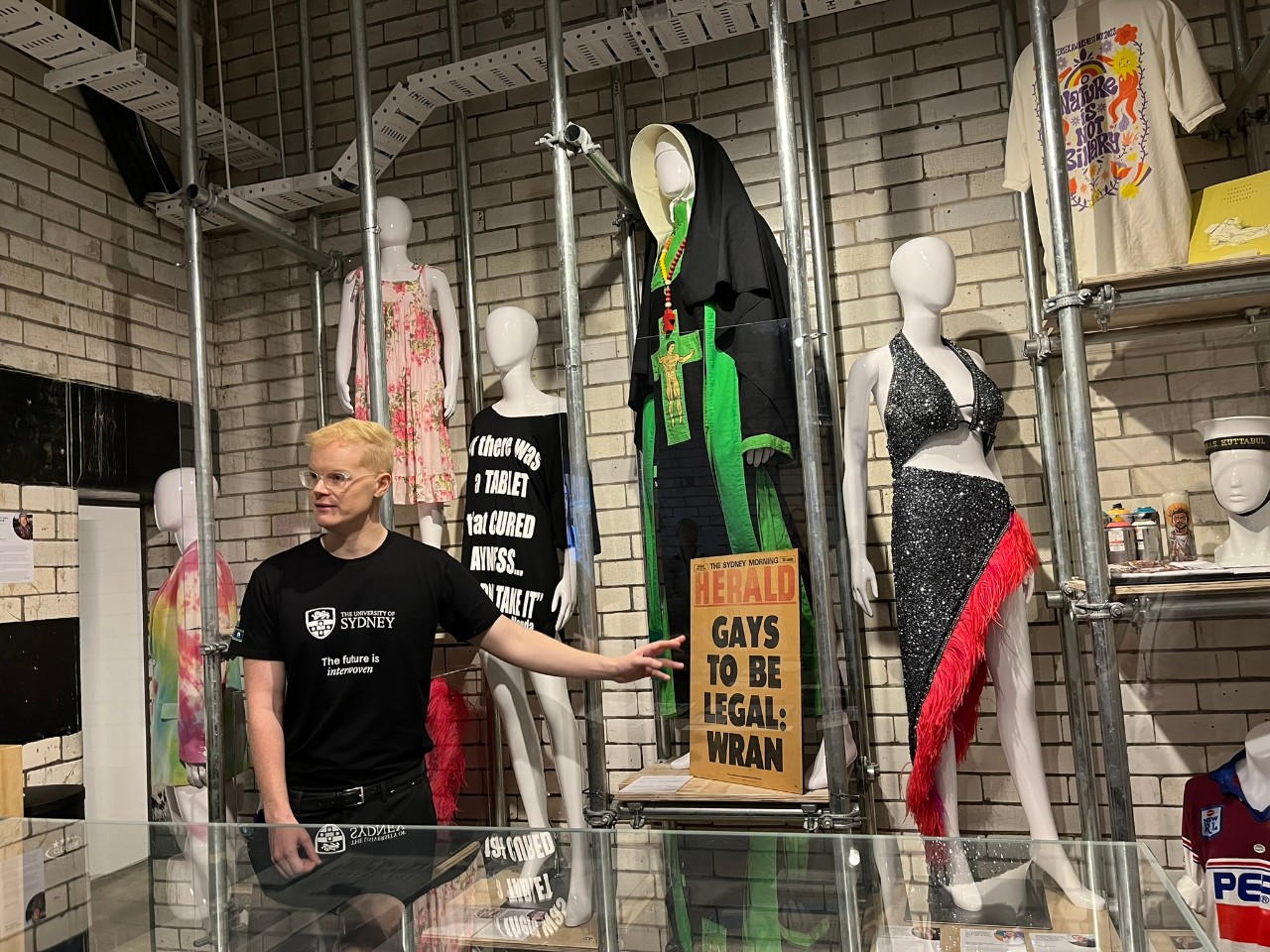 A photograph showing a white man with blonde hair and glasses, wearing a black tshirt with the University of Sydney logo, standing in front of an exhibition display. The display features a cover spread from an historical edition of the Sydney Morning Herald newspaper with the text "GAYS TO BE LEGAL: WRAN", and several mannequins wearing costumes of significance to the Sydney LGBTQIA+ community. These include a black t-shirt emblazened with white text reading "If there was a tablet that cured gayness... I wouldn't take it - Magda Szubanski", a costume similar to a Catholic nun's habit donated by protest and performance art group the Sisters of Perpetual Indulgence, and a sequinned costume worn by drag queen Courtney Act.